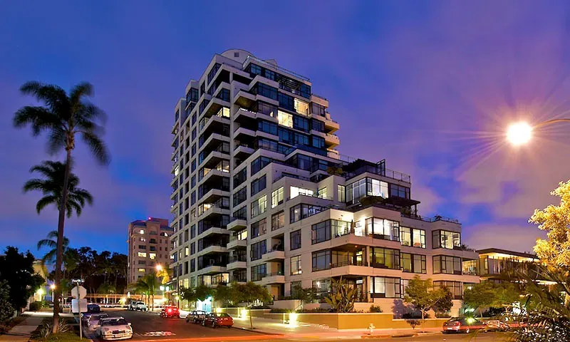 Modern multi-story residential building lit at dusk with palm trees and landscaping visible in twilight setting.
